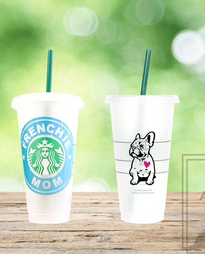 May include: Two white reusable coffee cups with lids and green straws. The cup on the left has a blue background with the text "Frenchie Mom" and a green Starbucks logo. The cup on the right has a black and white illustration of a French bulldog with a pink heart on its chest.