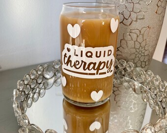 May include: A glass mug with brown liquid and white hearts printed on it. The mug has the text "Liquid therapy" printed on it.
