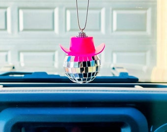 May include: A pink cowboy hat with a silver disco ball hanging from a rearview mirror in a car.