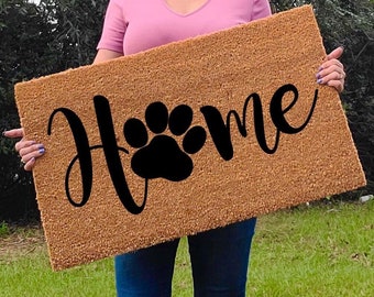 May include: A rectangular doormat with the word "Home" in a cursive font and a black paw print design. The mat is a natural brown color and is held by a person in a pink shirt and blue jeans.