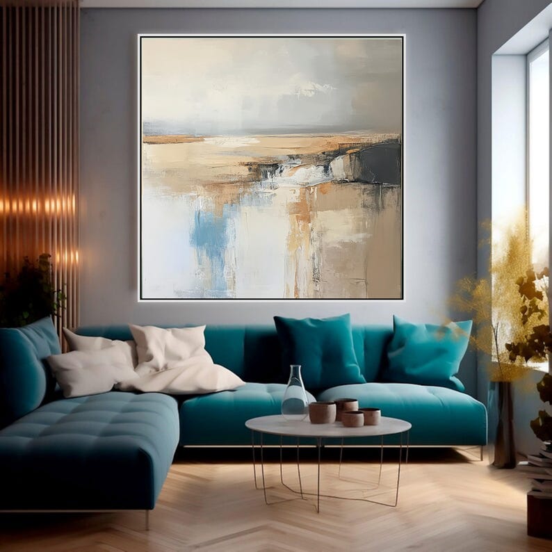 May include: Large abstract landscape painting in a black frame. The artwork features a muted color palette of beige, brown, blue, and white, creating a serene and reflective scene. The painting hangs above a teal sofa with white and teal pillows.