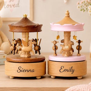 May include: Two wooden carousel music boxes with rotating horses. The left carousel has a dark brown roof and base with the name Sienna engraved. The right carousel has a light pink roof and base with the name Emily engraved. Both feature gold accents.