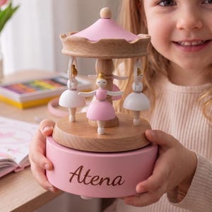 May include: A wooden carousel music box featuring three ballerina figures in white and pink dresses. The base is painted light pink with the name Atena written in dark script. The top canopy is pink, and the central structure is natural wood.