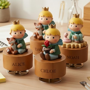 May include: Four wooden music boxes featuring a cartoon prince with a gold crown, green jacket, and blue scarf. Each box has a different design, including a fox, a rose, and a snake. The names "ALICE", "CHLOE", "DAV" and "NOAH" are engraved on the boxes.