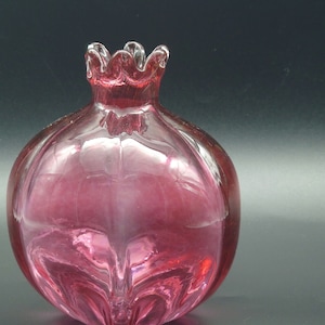 May include: A pink glass vase shaped like a pomegranate. The vase has a textured surface and a clear glass top.