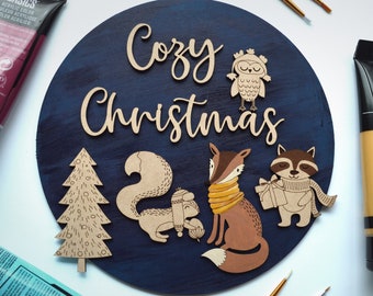 May include: A blue wooden round sign with the words "Cozy Christmas" in brown lettering. The sign features a brown owl, a fox, a raccoon, a squirrel, and a Christmas tree, all cut out of wood.