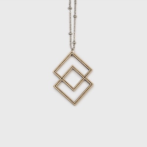 May include: A geometric pendant necklace featuring two overlapping square shapes. The pendant is made of light-colored wood with dark brown edges, suspended from a silver-colored chain with small, round beads. The necklace hangs against a white background.