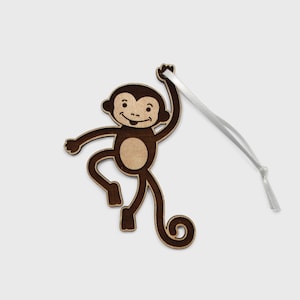 Puede incluir: Un adorno de mono de madera con una mancha marrón y una cinta blanca. El mono está levantando el brazo y tiene la cola enroscada.