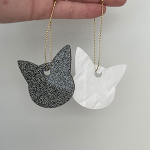 May include: Two cat-shaped ornaments, one black with glitter and one white with a pearlescent finish. Both have gold string for hanging.