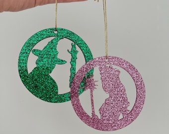 Puede incluir: Dos adornos redondos cubiertos de purpurina con la silueta de la Bruja Mala del Oeste y Glinda la Buena Bruja. El adorno de la Bruja Mala es verde y el de Glinda es rosa.