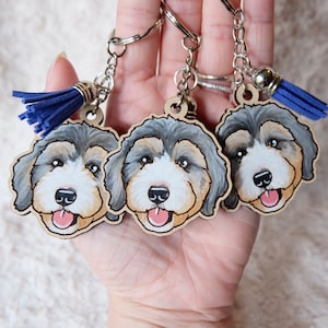 May include: Three wooden keychains featuring hand-painted portraits of dogs. Each keychain has a blue tassel attached to the metal ring.