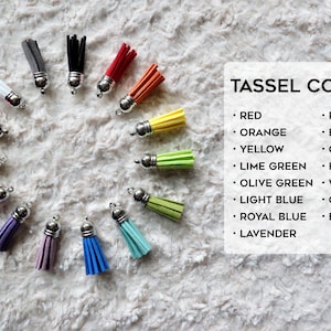 May include: A variety of colors of tassel keychains, including red, orange, yellow, lime green, olive green, light blue, royal blue, lavender, purple, baby pink, coral, hot pink, white, grey, and black. The tassels are arranged in a circle on a white fuzzy surface. The text "TASSEL COLORS" is written in black at the top of the image.