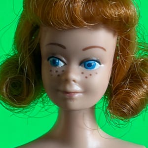 May include: A doll head with shoulder-length ginger hair styled in curls. The doll features a light skin tone, bright blue eyes, brown eyebrows, and small brown freckles across the nose and cheeks. The expression is neutral with a closed mouth.