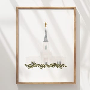 Può includere: Una stampa ad acquerello incorniciata raffigurante un tempio bianco con un'alta guglia grigia sormontata da una piccola figura dorata. Una ghirlanda orizzontale di foglie verdi è posizionata alla base dell'edificio, centrata su sfondo bianco.