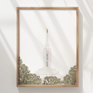 May include: A framed art print featuring a white temple building with a central spire topped by a small gold statue. The structure is framed by green foliage at the base, all enclosed within a light-toned wooden frame against a white background.