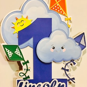 May include: A blue number one cake topper with a smiling sun, clouds, and kites. The name "Lincoln" is written in white script at the bottom. The kites are orange, green, and blue. The topper is supported by wooden dowels.