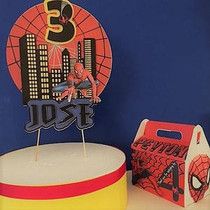 May include: A red and blue Spiderman themed cake topper with a cityscape backdrop, the number 3, and the name "JOSE". A red and blue Spiderman themed favour box with the name "PEYTON" and the number 4.