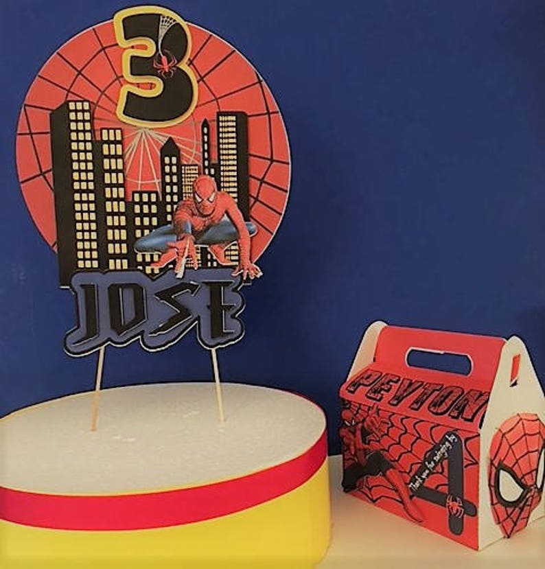 May include: A red and blue Spiderman themed cake topper with a cityscape backdrop, the number 3, and the name "JOSE". A red and blue Spiderman themed favour box with the name "PEYTON" and the number 4.