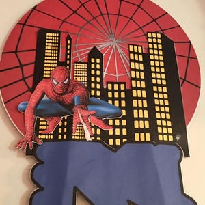 May include: A red, black and blue Spiderman cake topper with a web design and a cityscape backdrop. The topper features a Spiderman figure in a crouching pose, ready to swing into action.