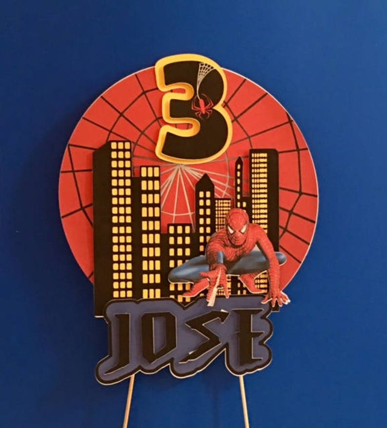May include: A red, white and black cake topper featuring a Spiderman character, a cityscape, a web and the number 3. The name "JOSE" is written in black letters on a blue background.