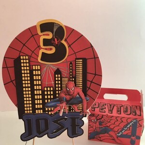 May include: Spider-Man themed birthday party decorations with a red and black cake topper featuring a cityscape, a number 3, and Spider-Man. The cake topper is on a wooden stick. A red and black favour box with Spider-Man and the name Peyton is also included. The favour box has the text "Thank you for making my day!"