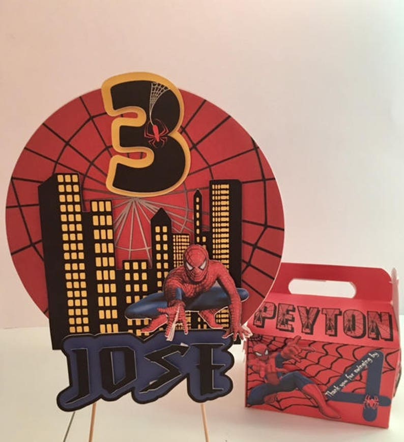 May include: Spider-Man themed birthday party decorations with a red and black cake topper featuring a cityscape, a number 3, and Spider-Man. The cake topper is on a wooden stick. A red and black favour box with Spider-Man and the name Peyton is also included. The favour box has the text "Thank you for making my day!"