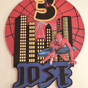 May include: A red, white and blue cake topper featuring a Spiderman figure, a cityscape, a spiderweb and the number 3. The name "JOSE" is written in black letters on a blue background.