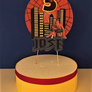 May include: A red, black and yellow Spiderman cake topper with a web design and the number 3. The topper also includes the name "JOSE" in black letters.