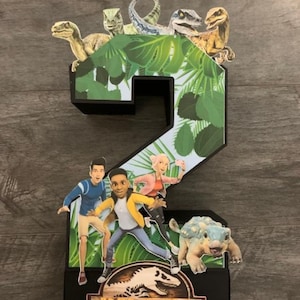 Puede incluir: Un adorno para pastel de número dos negro y verde con temática de selva. El adorno presenta dinosaurios, una tortuga y personajes del programa de televisión Jurassic World Camp Cretaceous. El adorno también incluye el nombre "KAYDEN".