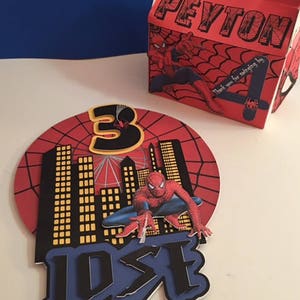 May include: Red and blue Spiderman cake topper featuring a Spiderman figure, city skyline, and the name "JOSE" in black letters. The topper is set against a red and black background with a spiderweb pattern. The text "Thank you for coming by" is on a red box with the name "PEYTON" in white letters.