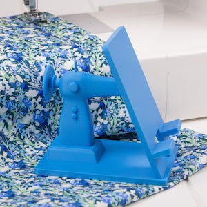 May include: A bright blue sewing machine-shaped phone holder sits on a floral patterned fabric. The holder is next to a sewing machine, with the needle positioned over the fabric. The fabric has a blue and white floral design.