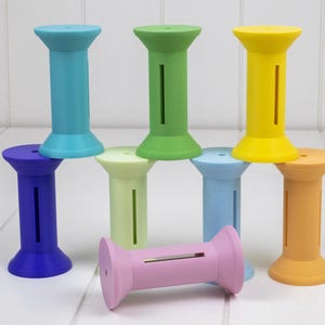 May include: A collection of colorful, spool-shaped objects in various shades of blue, green, yellow, orange, purple, and pink. Each spool has a cylindrical body with a slot cut down the side and a flared top and bottom. The objects are arranged in a playful stack.