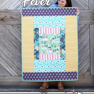 May include: A colorful quilt with a central rectangular design featuring floral and geometric patterns, and the text "Cabin Fever". The quilt has a yellow border and a purple patterned edge. The quilt is held up by a person.