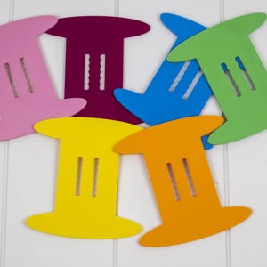 May include: A collection of colorful, wooden spool-shaped cutouts in pink, purple, blue, green, yellow, and orange. Each cutout has two vertical slots. The cutouts are arranged on a white background.