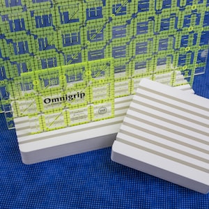 May include: Clear Omnigrip quilting rulers with grid markings and a white and gray striped cutting mat. The rulers have yellow and green accents. The text "Omnigrip" is visible on one ruler. The background is a blue fabric.
