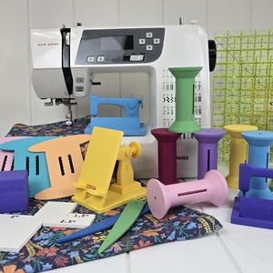 May include: A white sewing machine is surrounded by colorful sewing accessories. There are various spool holders, sewing machine-shaped objects, and a clear grid ruler. The accessories are in shades of pink, yellow, green, blue, and purple. A floral fabric is also present.