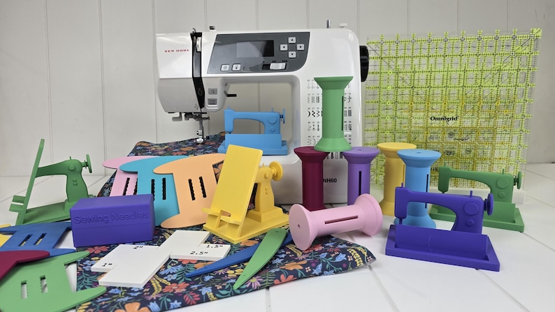 May include: A white sewing machine is surrounded by colorful sewing accessories. There are various spool holders, sewing machine-shaped objects, and a clear grid ruler. The accessories are in shades of pink, yellow, green, blue, and purple. A floral fabric is also present.