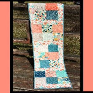 May include: A table runner with a patchwork design featuring squares of pink, blue, and white fabric. The runner is called "Macarons" and is designed by Tricia Lynn Maloney for Orphan Quilt Designs.
