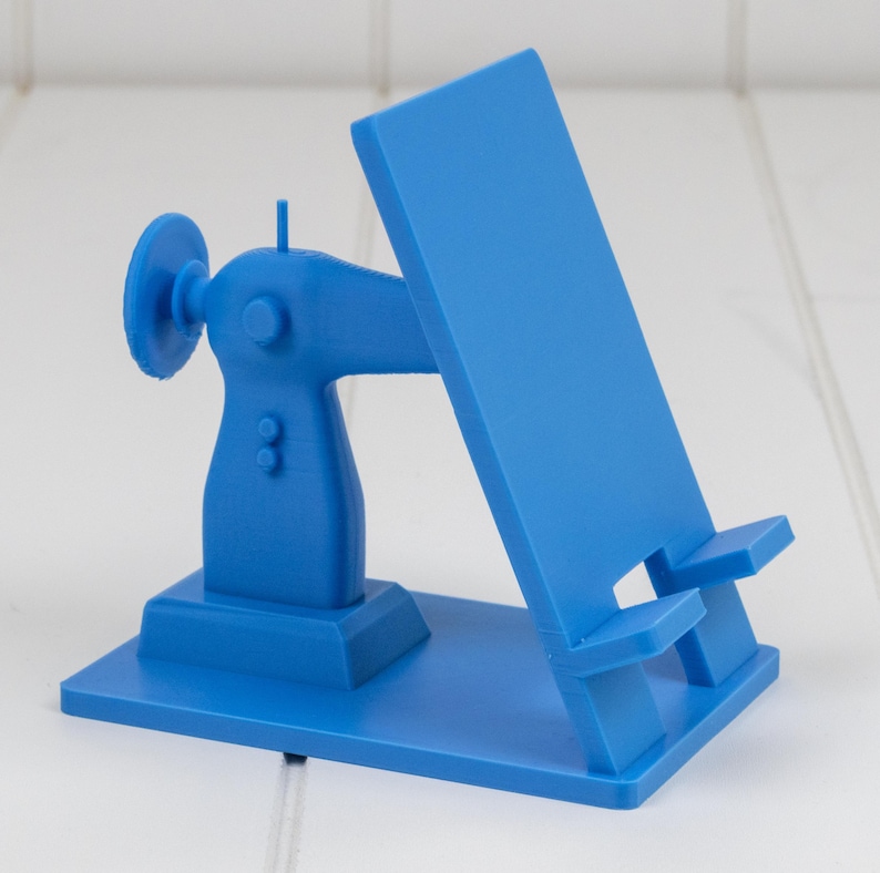 May include: A blue phone stand shaped like a vintage sewing machine. The stand has a vertical back and a base with a sewing machine design. The stand is made of plastic and is designed to hold a phone or tablet.