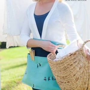 May include: A woman wearing a white cardigan and a teal blue apron with a black and white design of birds. She is holding a wicker basket filled with white fabric. The apron has a black belt and two pockets on the sides. The text "First Blush Clothespin Apron" is at the bottom of the image.