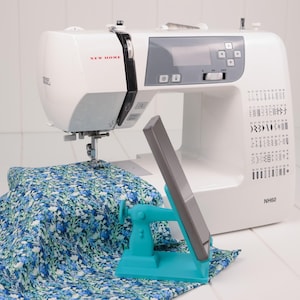 May include: A white New Home sewing machine with a gray control panel and a blue and white floral fabric. A teal phone holder is in front of the machine. The model number NH60 is visible.