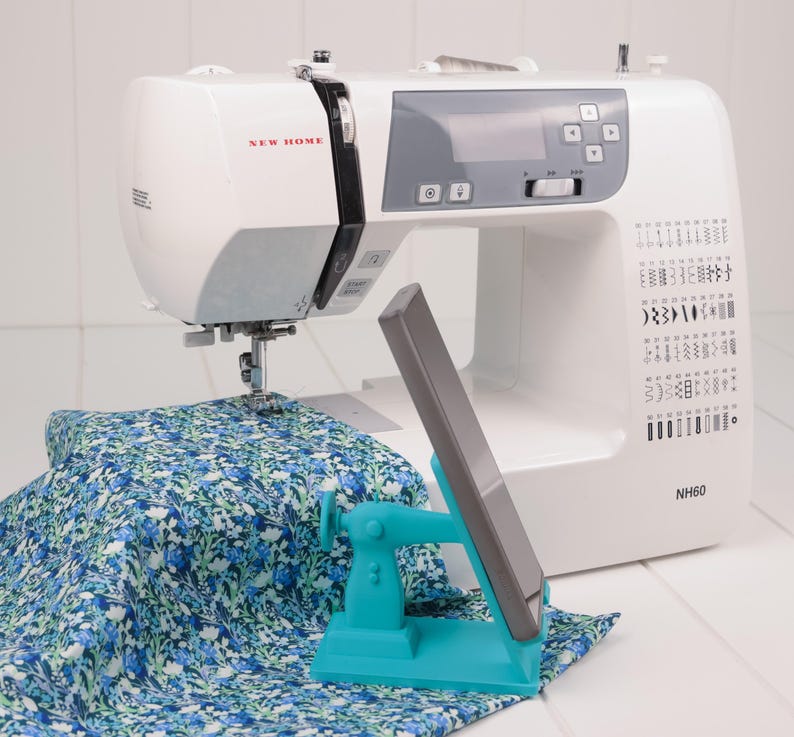 May include: A white New Home sewing machine with a gray control panel and a blue and white floral fabric. A teal phone holder is in front of the machine. The model number NH60 is visible.
