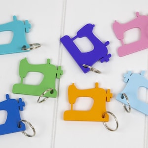 May include: A collection of colorful sewing machine keychains in shades of blue, green, purple, pink, and orange. Each keychain features a metal ring for attaching to keys or bags. The keychains are made of a solid material.