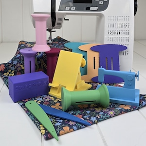 May include: A collection of colorful sewing accessories, including thread spool holders, a phone stand, and a miniature sewing machine. The accessories are in various colors, such as pink, purple, yellow, green, and blue. A purple box with the text "Sewing Needles" is also present.