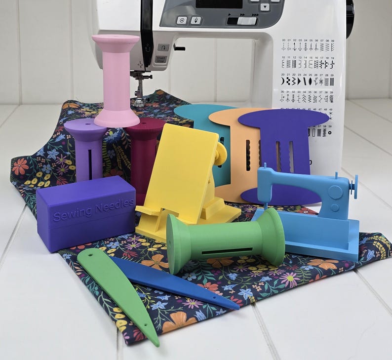 May include: A collection of colorful sewing accessories, including thread spool holders, a phone stand, and a miniature sewing machine. The accessories are in various colors, such as pink, purple, yellow, green, and blue. A purple box with the text "Sewing Needles" is also present.