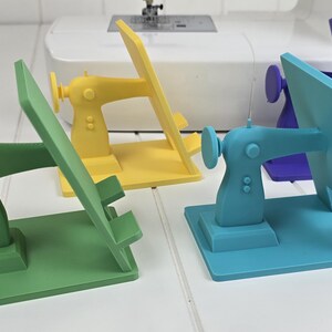 May include: Four colorful sewing machine-shaped phone stands in green, yellow, teal, and purple. Each stand has a rectangular base and a back panel to hold a phone. A white sewing machine is in the background.