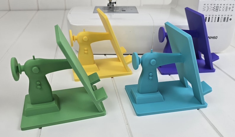 May include: Four colorful sewing machine-shaped phone stands in green, yellow, teal, and purple. Each stand has a rectangular base and a back panel to hold a phone. A white sewing machine is in the background.