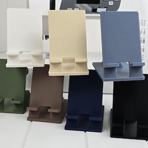 May include: A collection of seven phone stands in various colors: white, beige, gray-blue, green, brown, navy blue, and black. Each stand has a rectangular back and a base with a slot to hold a phone.