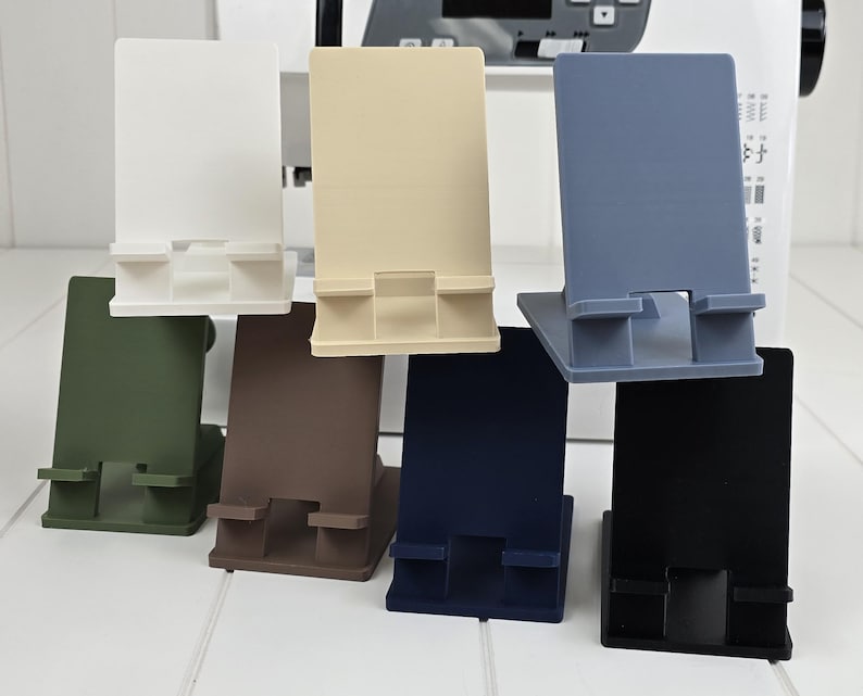 May include: A collection of seven phone stands in various colors: white, beige, gray-blue, green, brown, navy blue, and black. Each stand has a rectangular back and a base with a slot to hold a phone.