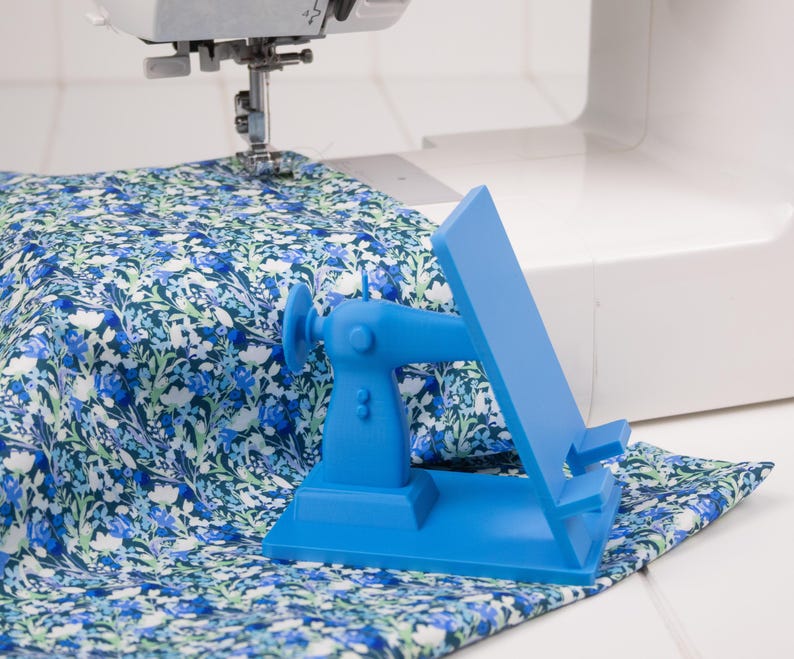 May include: A bright blue sewing machine-shaped phone holder sits on a floral patterned fabric. The holder is next to a sewing machine, with the needle positioned over the fabric. The fabric has a blue and white floral design.